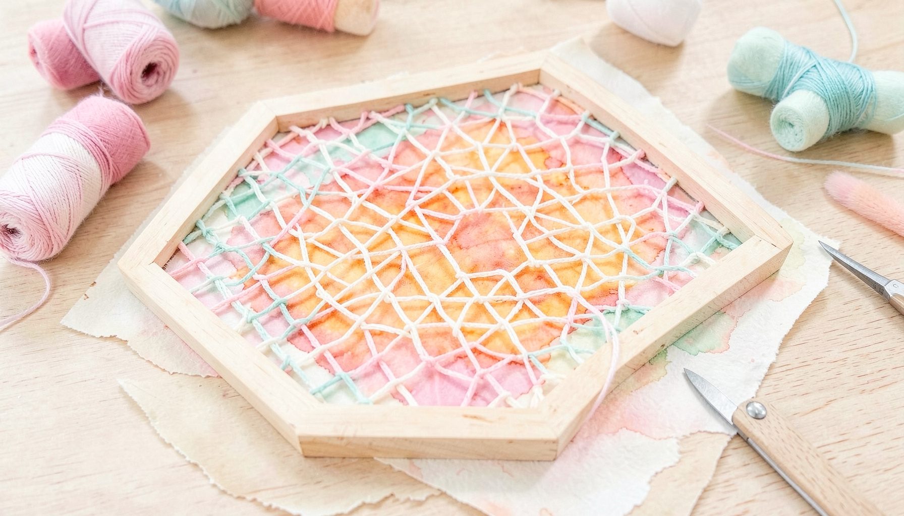 Weiche Aquarellmalerei in sanften Pastelltönen: ein handgefertigter Webrahmen aus hellem Holz mit begonnenem Gewebe in Rosa, Creme und Mintgrün, umgeben von Garnrollen und einer kleinen Schere auf einem hellen Holztisch.