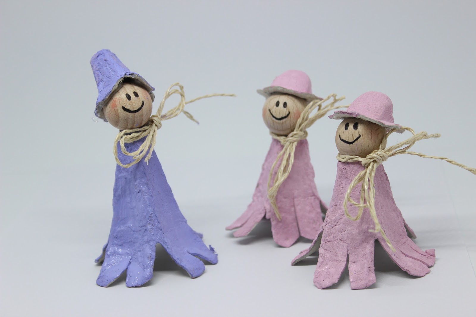 Drei kleine handgefertigte Figuren mit Holzkugelköpfen und bemalten Eierkartonkörpern in sanftem Blau und Rosa, mit Bindfaden verziert, auf einer hellen Fläche arrangiert